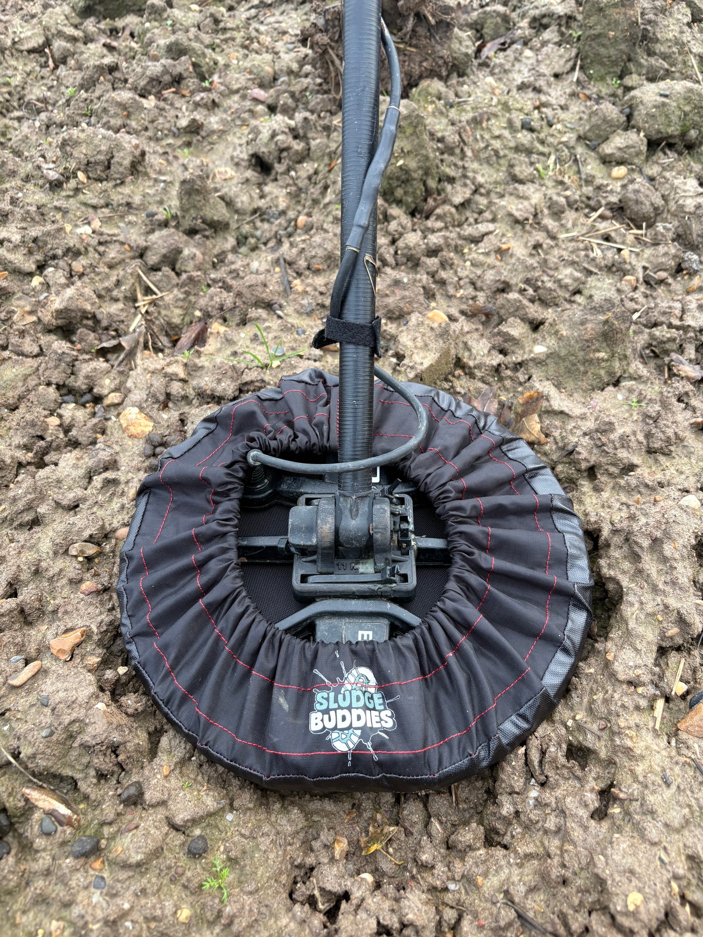 Metal detector coil fitted with a Sludge Buddies coil cover, shown in a muddy field during real-world use.