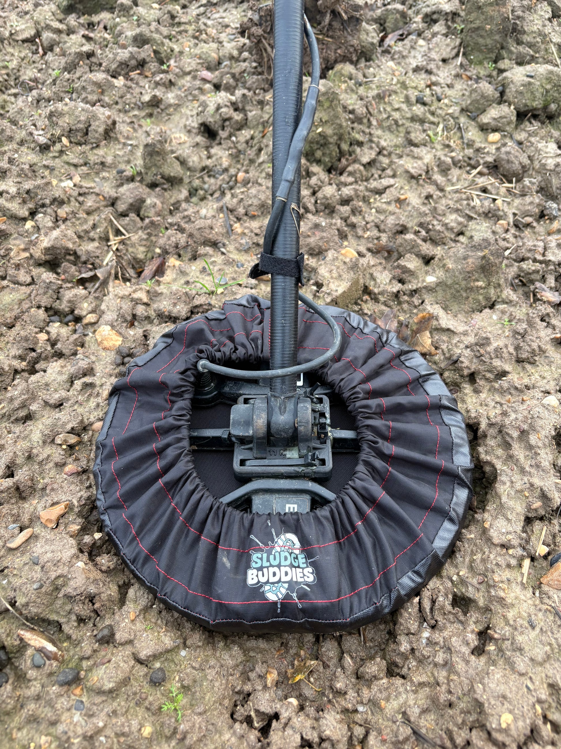 Metal detector coil fitted with a Sludge Buddies coil cover, shown in a muddy field during real-world use.